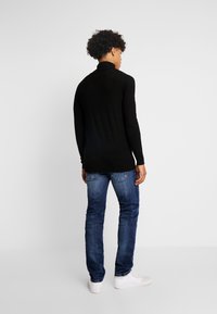 Crni turtleneck pulover dugih rukava uparen s plavim farmerkama i bijelim tenisicama, prikazan s leđa, naglašavajući teksturu i kroj.