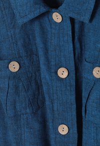 Blå linneskjorta med krage, fyra knappar och två bröstfickor med träknappar. Texturerat tyg med sömdetaljer.