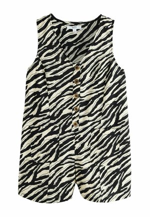 Frau trägt ärmellosen schwarz-weißen Zebra-Print-Overall mit Knöpfen, steht neben einer strukturierten Wand und einem Geländer mit Grün im Hintergrund.