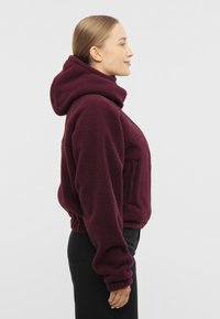 Burgundinvärgad fleecehuva med hög krage, elastiska ärmmuddar och en framficka. Mjuk textur och avslappnad passform. Stylad med svarta byxor.