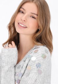 Jasnoszary, puszysty sweter z długimi rękawami, zdobiony okrągłymi, iryzującymi elementami po jednej stronie. Miękka struktura i dekolt w serek.