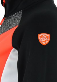 Veste de sport noire et orange présentant un panneau gris texturé, un écusson orange sur l'épaule et des accents de couleur vifs.