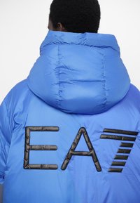 Zilā, polsterētā jaka ar lielu melnu "EA7" logo aizmugurē. Ar augstu apkakli un gludu, spīdīgu tekstūru.