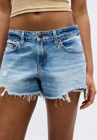 Pantalones cortos de mezclilla azul claro deshilachados, usados por una persona de piel marrón media y top corto beige, sobre un fondo blanco liso.