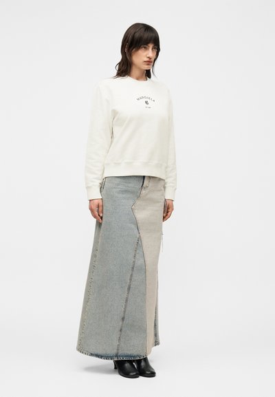 Γυναίκα με σκούρα μαλλιά που φοράει λευκό φούτερ Margiela, μακριά δίχρωμη τζιν φούστα και μαύρα μποτάκια, στέκεται μπροστά σε απλό φόντο.