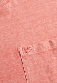 Gros plan sur un tissu rose corail avec une poche cousue affichant un petit logo brodé en fil assorti sur le bord de la poche.