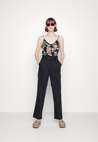Camisola preta floral com alças finas, combinada com calças pretas de cintura alta e sandálias cinzentas claras. A modelo está a usar óculos de sol escuros.