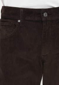 Pantalones de pana marrones con acanalado vertical, bolsillos delanteros y cierre de botón metálico en la cintura. La superficie tiene una textura suave.