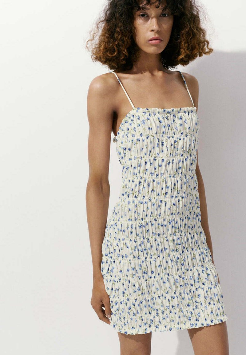 Femme aux cheveux bouclés portant une robe blanche sans manches à imprimé floral bleu, debout devant un fond clair uni.