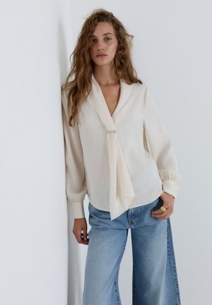Frau mit welligem Haar, die eine cremefarbene Langarmbluse mit Bindedetail und hellblaue, hoch taillierte Jeans trägt, stehend vor einer weißen Wand.