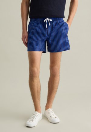 Costume da bagno a righe blu e nere con cintura elastica e lacci bianchi. Include tasche laterali e un piccolo logo decorativo.
