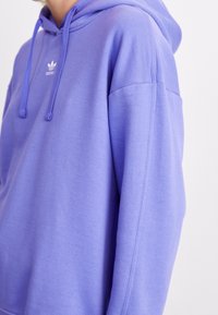 Levandulová mikina s kapucí vyrobená z měkké tkaniny a s volným střihem. Má bílé logo na hrudi a šňůrkové detaily na kapuci.