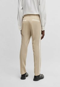 Beige maßgeschneiderte Hosen mit seitlichem Streifen, glattem Stoff und zwei Gesäßtaschen. Getragen mit einem weißen Hemd und schwarzen Schuhen.