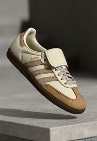 Kremfargede og beige lærsneakers med tre hvite striper, en polstret tunge og teksturert brun såle. "SAMBA"-etikett på siden.