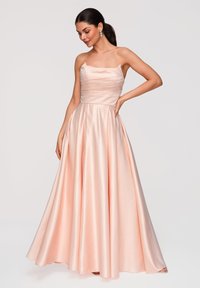 Femme aux cheveux foncés portant une robe bustier en satin rose avec un corsage plissé, debout, une main sur la taille, sur un fond clair uni.
