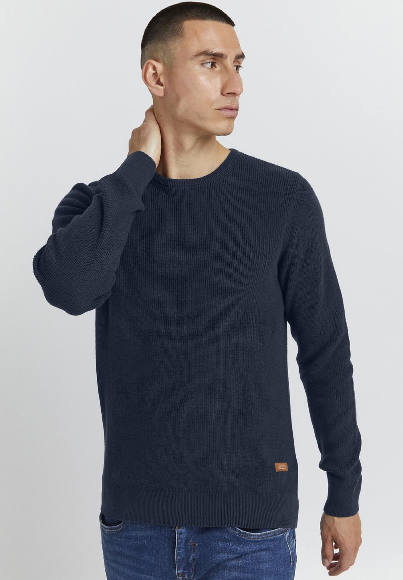 Pull en maille marine avec texture côtelée, col rond et manches longues. Présente une petite étiquette logotée près de l'ourlet et un design ajusté.