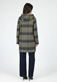 Imperméable à carreaux dans des tons vert olive, noir et bleu. Doté d'une capuche, d'une coupe mi-longue et de manches longues. Associé à un jean bleu foncé.