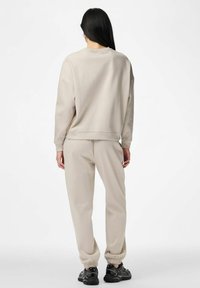 Moteris su ilgais tamsiais plaukais, vilkinti smėlio spalvos džemperį ir laisvalaikio kelnes, juodi sportbačiai, stovinti nugara į baltą foną.