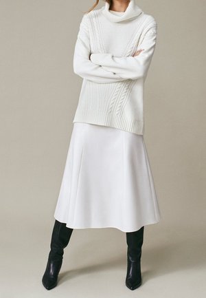 Person wearing a white cable-knit turtleneck sweater, white midi skirt, and black knee-high boots, standing with arms crossed.