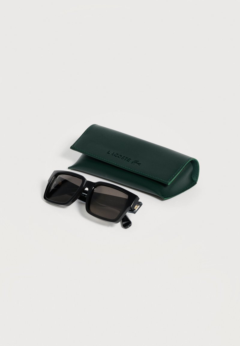 Occhiali da sole rettangolari neri con lenti scure accanto a una custodia per occhiali da sole Lacoste verde scuro su una superficie bianca.