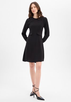 Femme debout portant une robe noire à manches longues, longueur genoux, avec des détails à rabats à la taille et des chaussures noires à talons hauts et bout pointu.