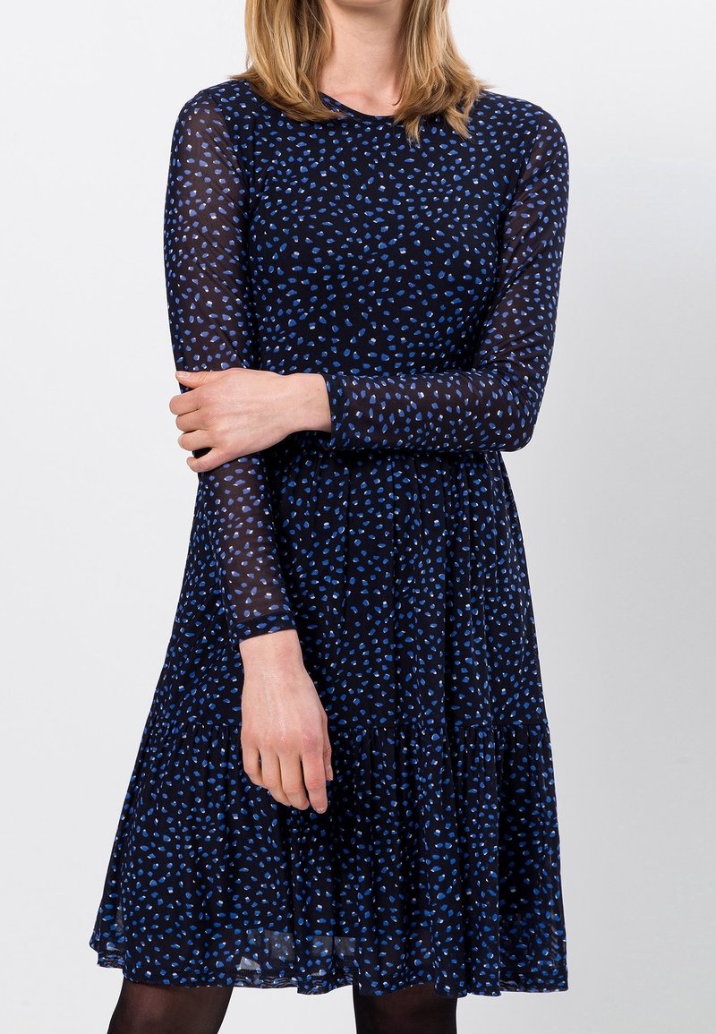 Robe bleu marine à manches longues avec des pois bleu clair dispersés, texture semi-transparente, corsage ajusté et jupe à volants se terminant sous le genou.