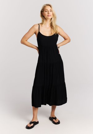 Blonde vrouw, staand met handen in de zij, draagt een mouwloze zwarte midikleding met lagen en zwarte slideschoenen, op een neutrale achtergrond.