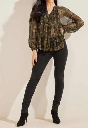 Femme portant un chemisier camouflage transparent à manches longues, pantalon noir près du corps et bottines noires à talons hauts, posant avec la main sur la hanche.