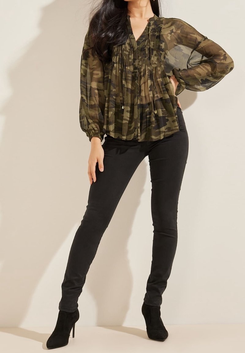 Femme portant un chemisier camouflage transparent à manches longues, pantalon noir près du corps et bottines noires à talons hauts, posant avec la main sur la hanche.