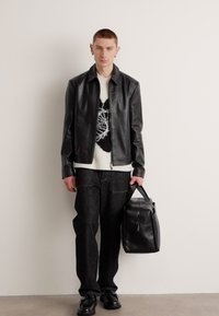 Giacca di pelle nera sopra un maglione bianco con disegno, pantaloni in denim scuri, scarpe nere e una borsa nera texturizzata. Design semplice e schema di colori monocromatico.