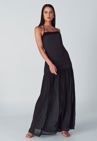 Schwarzes Maxikleid mit tailliertem Oberteil, dünnen Trägern und einem gefütterten, fließenden Rock. Glattes Material mit leichtem Glanz und minimalistischem Design.