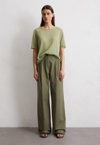 Light green, short-sleeve t-shirt paired with loose, high-waisted olive pants; model stands on a neutral-colored floor against a plain wall.