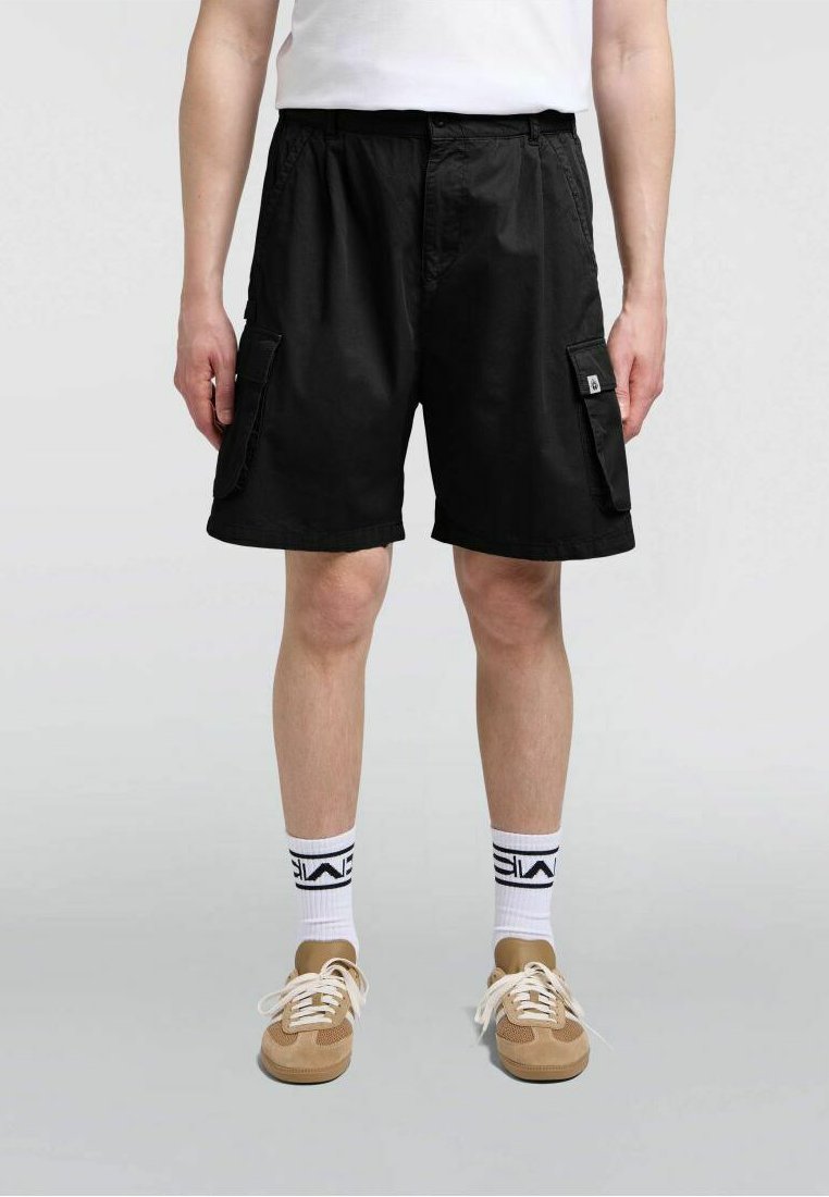 Pantaloncini cargo neri dal taglio ampio, con due tasche laterali e una vita con coulisse. Indossati con calzini bianchi e sneakers beige.