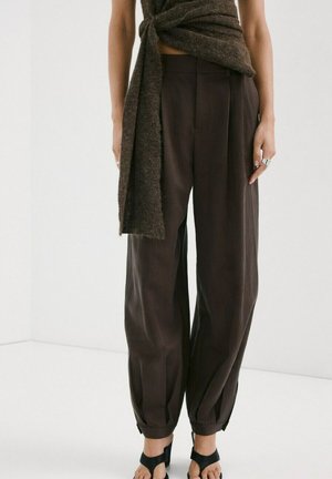 Persona que lleva pantalones anchos de cintura alta y color marrón oscuro con puños, tacones abiertos negros y una blusa sin mangas marrón texturizada atada en la cintura.