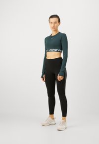 Ciemnozielony krótki top z długim rękawem z logo "Nike Pro", w połączeniu z czarnymi legginsami o wysokim stanie i jasnymi sneakersami. Gładka tkanina, dopasowany krój.