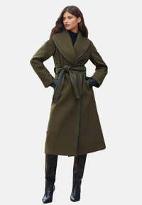 Alyvuogių žalios spalvos paltas su ilgu, apvyniojamu dizainu, plačiu apykaklė, juodu kraštu ir diržu. Dėvimas su juodais odiniais pirštiniais ir iki šlaunų siekiančiais batais.