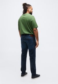 Άνδρας με ντραντλοκς που φοράει πράσινο μπλουζάκι, σκούρο μπλε τζιν και μαύρα αθλητικά παπούτσια, στέκεται με την πλάτη προς το μέρος μας μπροστά σε απλό λευκό φόντο.