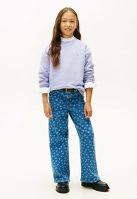 Lyse lilla sweatshirt med flæset halsudskæring og logo, parret med blå denimbukser med blomstermønster og sorte platform-sko.