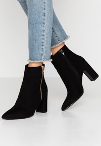 Botines de ante negro con punta afilada, cremallera lateral con herrajes dorados y tacón cuadrado. Combinados con jeans de dobladillo deshilachado.
