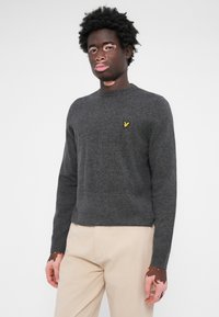 Pull en tricot gris foncé avec un col rond et des manches longues, présentant un petit logo jaune sur la poitrine gauche. La texture semble douce.