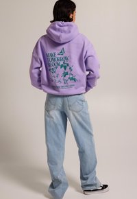 Sudadera con capucha de color lavanda que presenta un texto gráfico verde y un diseño floral en la parte trasera, combinada con jeans de pierna ancha azul claro y zapatillas negras.