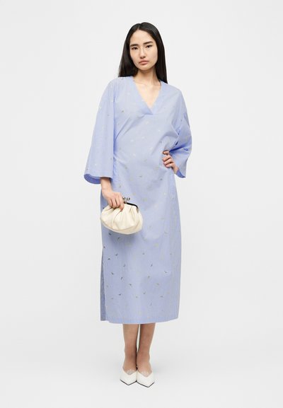 Μακρύ μπλε φόρεμα με ρίγες και μανίκια κιμονό, με μικρές χρυσές λεπτομέρειες. Το μοντέλο κρατάει μια κρεμ τσάντα, φοράει λευκά μυτερά παπούτσια.