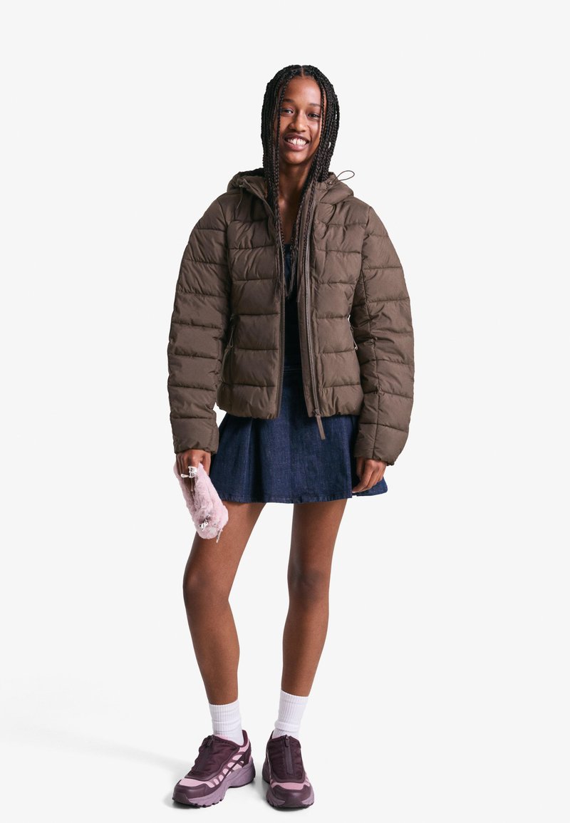 Rjava puhovka s kapuco, prešita zasnova in elastični manšeti. V kombinaciji z jeans krilom in vijoličnimi superge, drži roza torbico.