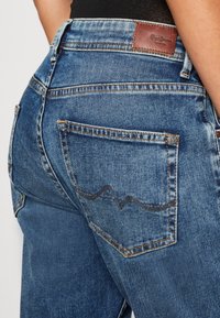Denim jeans with a blue wash, featuring a brown leather logo patch on the back waistband and distinctive stitching on the rear pockets.