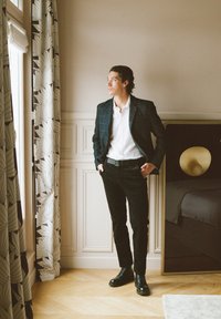 Blazer à motifs sombres sur une chemise blanche, un pantalon noir et des bottes noires. Se tenant près d'une fenêtre avec des rideaux légers et un mobilier moderne.