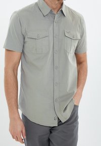 Camisa de manga corta abotonada en tejido gris claro. Presenta dos bolsillos en el pecho, un cuello con punta y un pequeño detalle de logo cerca del dobladillo.
