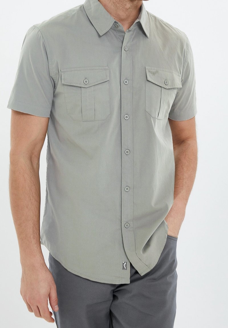 Camisa de manga corta abotonada en tejido gris claro. Presenta dos bolsillos en el pecho, un cuello con punta y un pequeño detalle de logo cerca del dobladillo.