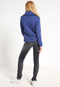 Sudadera azul con cuello de cowl en tejido suave, combinada con jeans negros y zapatillas blancas. El corte es relajado, con mangas largas y dobladillo acanalado.
