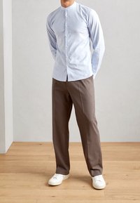 Camisa de manga larga azul claro con cuello mao, frontal abotonado, combinada con pantalones marrones y zapatillas blancas sobre suelo de madera.