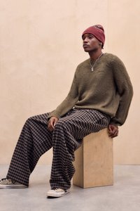 Olivenfarvet, pelsagtig sweater, brede brune og sorte ternede bukser, beige sneakers med sorte snørebånd og en rød strikket hue. Sidder på en træterning.